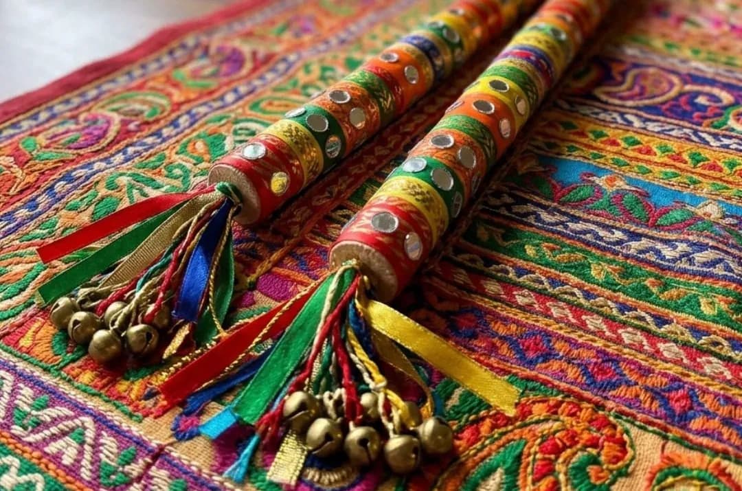 Two newspaper dandiya sticks wrapped in bright tape with mirror stickers, ribbons and bells, on an embroidered cloth