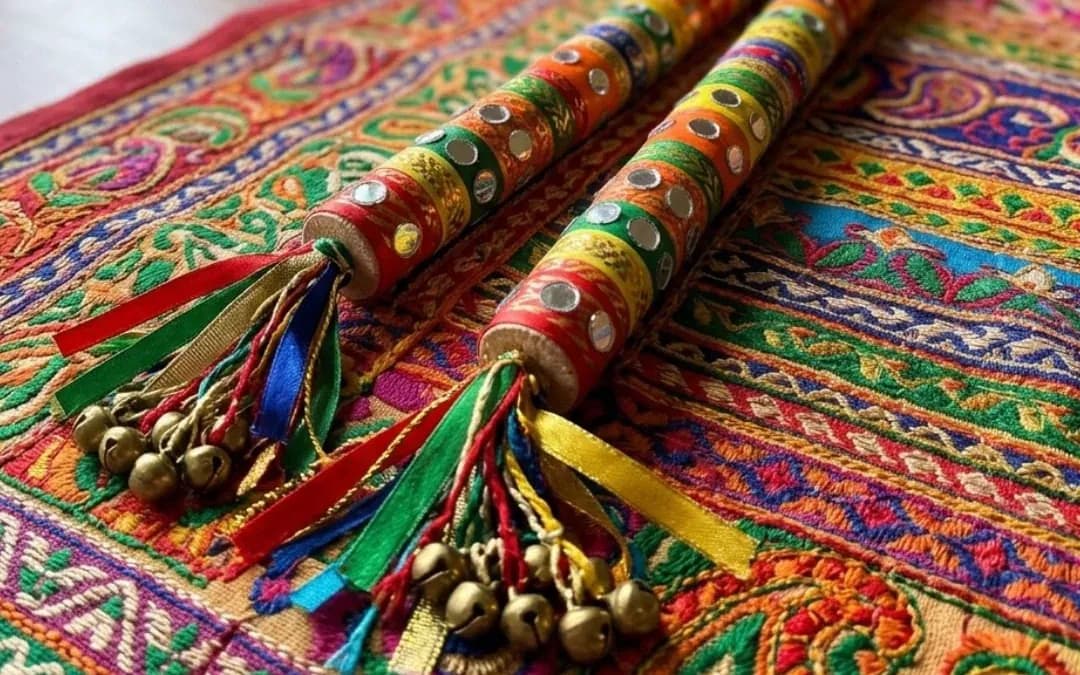 Two newspaper dandiya sticks wrapped in bright tape with mirror stickers, ribbons and bells, on an embroidered cloth